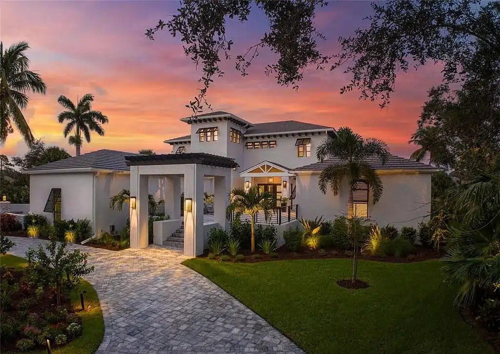 Two-story white Southwest Florida home with lit entry and paver driveway at dusk